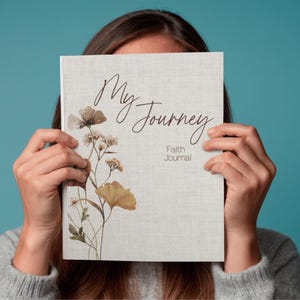 May include: A journal titled "My Journey" with the words "Faith Journal" below. The cover is light grey with a floral design in brown and yellow. The journal is held in front of a person.