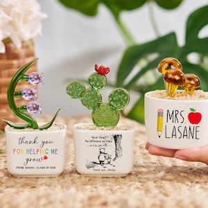 Pode incluir: Três pequenos vasos de cerâmica branca com esculturas de plantas de vidro. Um vaso tem um lírio-do-vale verde, outro um cacto verde e o terceiro esculturas de cogumelos. O texto nos vasos diz "Obrigado por me ajudar a crescer", "Nunca se esqueça da diferença que você faz" e "MRS. LASANE".