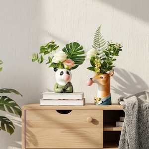 May include: Two decorative ceramic vases, one a panda and the other a deer, each filled with flowers. The panda vase is white with black ears and a green sweater. The deer vase is brown with a blue jacket. Both have pink bubblegum bubbles.