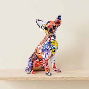 Peut inclure: Figurine de Chihuahua colorée avec un design abstrait vibrant. Le chien est assis, les oreilles dressées. L'œuvre présente un mélange de rouge, jaune, bleu et blanc, créant un effet visuel saisissant. La figurine est exposée sur une étagère en bois.