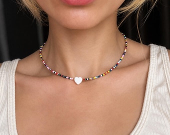 Puede incluir: Un collar de cuentas con un colgante de corazón blanco. El collar presenta pequeñas cuentas de colores en una variedad de colores, incluyendo rosa, amarillo, azul y negro. El colgante de corazón está centrado.