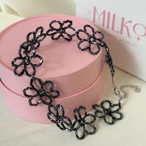 May include: A necklace constructed from dark grey, cylindrical glass beads arranged into a series of connected flower shapes. The piece features a silver-toned metal lobster clasp and an adjustable chain extension with a small circular metal tag.