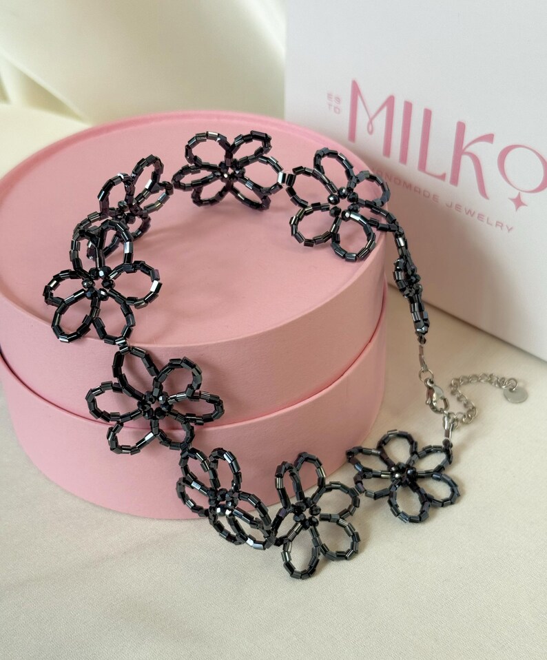 May include: A necklace constructed from dark grey, cylindrical glass beads arranged into a series of connected flower shapes. The piece features a silver-toned metal lobster clasp and an adjustable chain extension with a small circular metal tag.