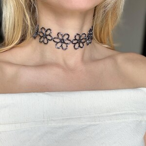 May include: A dark grey beaded choker necklace featuring a repeating floral pattern. The necklace is constructed from small, reflective metallic-toned beads arranged into interconnected flower shapes that sit closely around the neck.