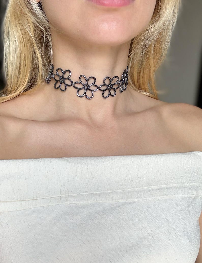 May include: A dark grey beaded choker necklace featuring a repeating floral pattern. The necklace is constructed from small, reflective metallic-toned beads arranged into interconnected flower shapes that sit closely around the neck.