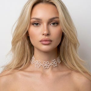 May include: A white floral choker necklace. The necklace features a series of delicate, openwork flower designs. The choker is worn around the neck and is a stylish accessory.