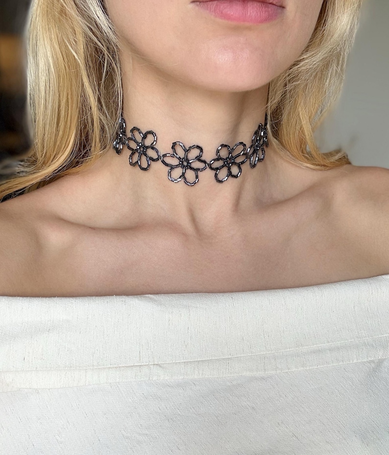 May include: A dark grey beaded choker necklace featuring a repeating pattern of interconnected flower shapes. The necklace is constructed from small, reflective, metallic-toned beads arranged to form individual floral outlines around the neck.