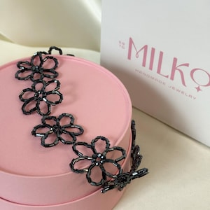 May include: A black beaded necklace featuring a series of connected flower-shaped links made from small, dark, cylindrical beads. The necklace is draped over a round, light pink container next to a white box with the text MILKQ HANDMADE JEWELRY.
