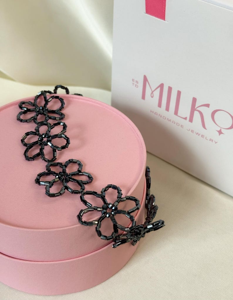 May include: A black beaded necklace featuring a series of connected flower-shaped links made from small, dark, cylindrical beads. The necklace is draped over a round, light pink container next to a white box with the text MILKQ HANDMADE JEWELRY.