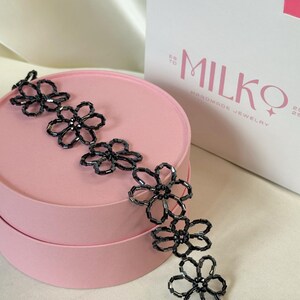 May include: A black beaded necklace featuring a series of connected flower-shaped motifs made from small, dark, cylindrical beads. The necklace is draped across a round, light pink box, with a white gift bag labeled MILKO HANDMADE JEWELRY nearby.