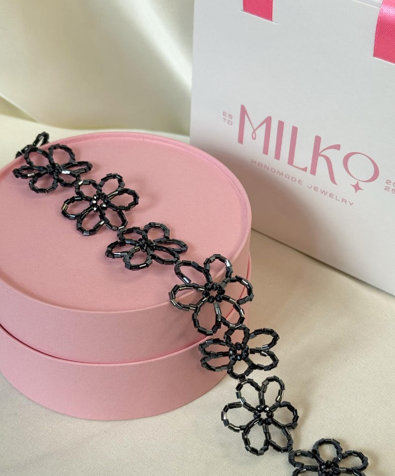 May include: A black beaded necklace featuring a series of connected flower-shaped motifs made from small, dark, cylindrical beads. The necklace is draped across a round, light pink box, with a white gift bag labeled MILKO HANDMADE JEWELRY nearby.