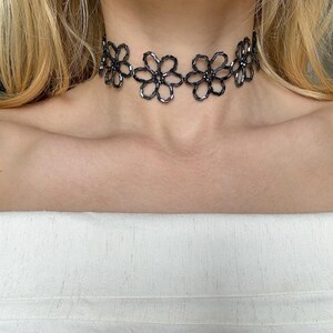 May include: A black beaded choker necklace featuring a repeating floral pattern. The necklace is constructed from small, dark, reflective cylindrical beads arranged into individual flower shapes that are linked together to form a continuous band.