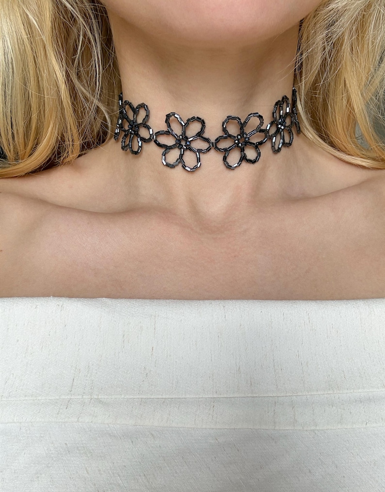May include: A black beaded choker necklace featuring a repeating floral pattern. The necklace is constructed from small, dark, reflective cylindrical beads arranged into individual flower shapes that are linked together to form a continuous band.