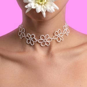 May include: A white beaded floral choker necklace. The necklace features a series of openwork flower designs. A white flower is held near the mouth. The background is a solid pink.