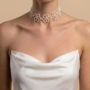 May include: A white floral choker necklace. The necklace features a series of delicate, openwork flower designs. The model is wearing a white satin top. The background is a solid, neutral color.