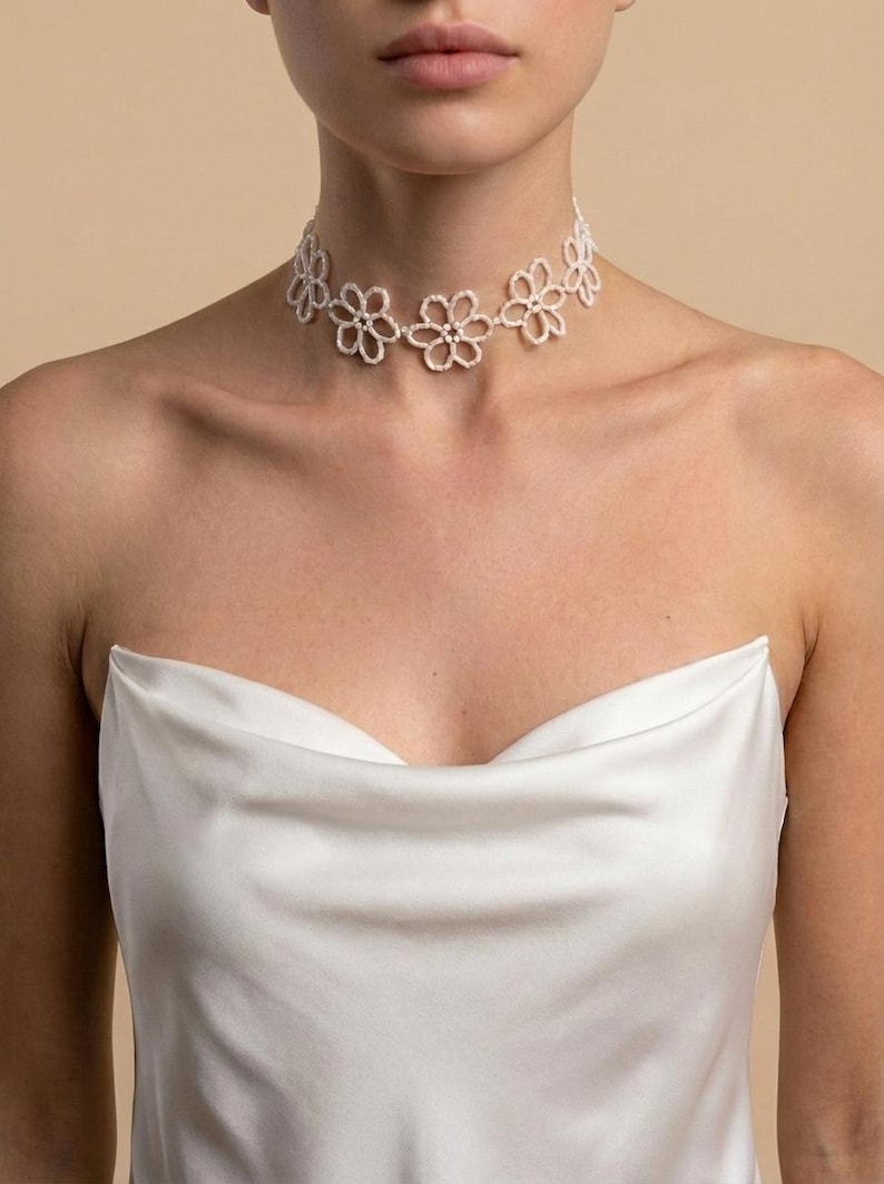 May include: A white floral choker necklace. The necklace features a series of delicate, openwork flower designs. The model is wearing a white satin top. The background is a solid, neutral color.