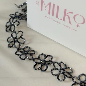 May include: A necklace composed of dark gray, metallic-finish beads arranged in a repeating flower pattern. The necklace is positioned next to a white box featuring the text "ESTD MILKQ HANDMADE JEWELRY" in pink and gray lettering.