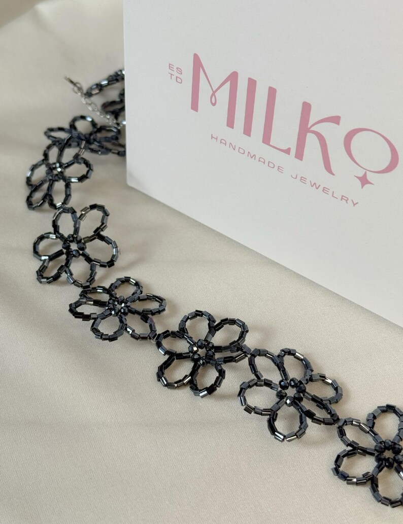 May include: A necklace composed of dark gray, metallic-finish beads arranged in a repeating flower pattern. The necklace is positioned next to a white box featuring the text "ESTD MILKQ HANDMADE JEWELRY" in pink and gray lettering.