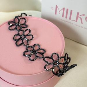 May include: A dark gray beaded necklace featuring a series of interconnected flower-shaped motifs. Each flower is constructed from small, cylindrical glass beads arranged in a circular pattern, creating a delicate and repeating floral design.