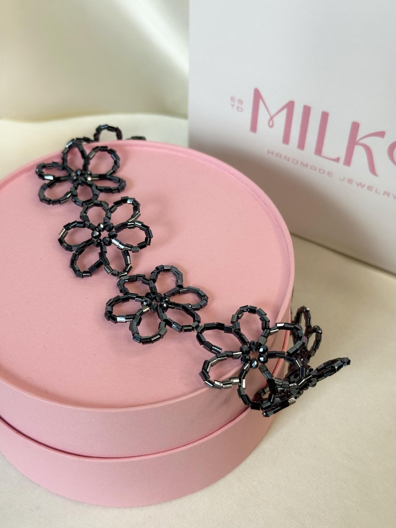 May include: A dark gray beaded necklace featuring a series of interconnected flower-shaped motifs. Each flower is constructed from small, cylindrical glass beads arranged in a circular pattern, creating a delicate and repeating floral design.