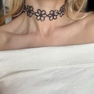 May include: A dark gray beaded choker necklace featuring a repeating pattern of interconnected flower shapes. The necklace is constructed from small, reflective cylindrical beads arranged to form the petals and centers of each floral motif.