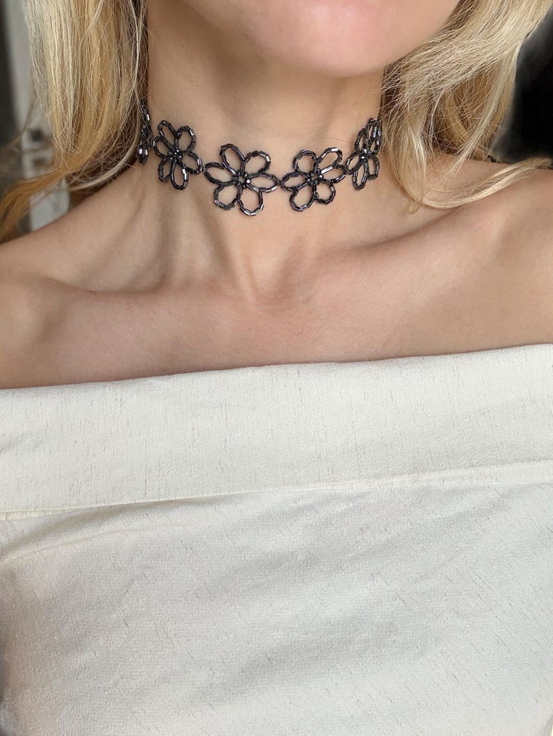 May include: A dark gray beaded choker necklace featuring a repeating pattern of interconnected flower shapes. The necklace is constructed from small, reflective cylindrical beads arranged to form the petals and centers of each floral motif.