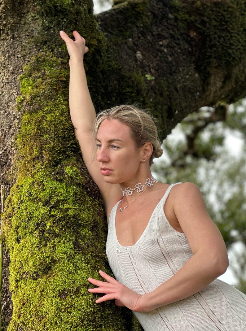 May include: A white floral choker necklace and matching pendant necklace. The model is wearing a white knit dress. The background features a moss-covered tree trunk.