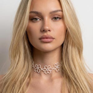 May include: A delicate, white floral choker necklace. The necklace features three open flower designs, each with a central bead. The choker is worn around the neck and is a light, airy accessory.