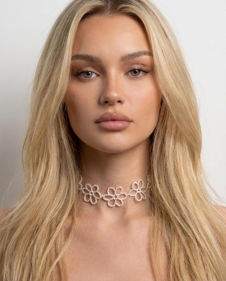 May include: A delicate, white floral choker necklace. The necklace features three open flower designs, each with a central bead. The choker is worn around the neck and is a light, airy accessory.