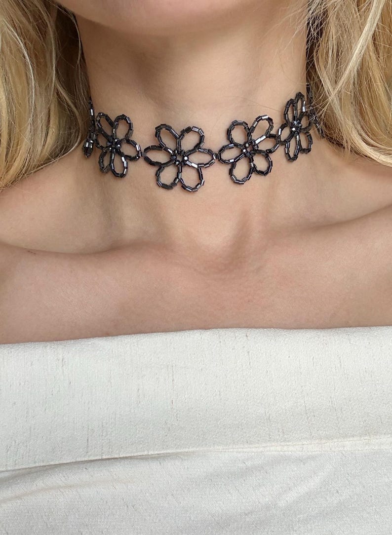 May include: A choker necklace crafted from dark gray, metallic-toned seed beads arranged in a repeating floral pattern. The individual flowers are linked together to form a continuous band that sits snugly around the neck, featuring a reflective finish.