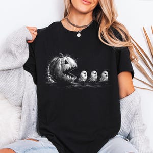 May include: A black short-sleeved t-shirt featuring a graphic print of a large, textured jack-o'-lantern with a wide, jagged mouth appearing to chase three small, white, round ghosts in a line across the centre of the chest.