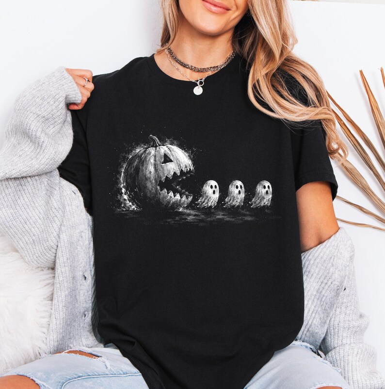 May include: A black short-sleeved t-shirt featuring a graphic print of a large, textured jack-o'-lantern with a wide, jagged mouth appearing to chase three small, white, round ghosts in a line across the centre of the chest.