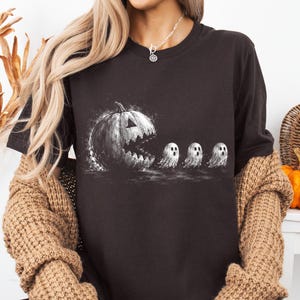 May include: A black short-sleeved t-shirt featuring a white graphic print of a large jack-o'-lantern pumpkin with a jagged mouth chasing three small ghosts in a line. The design has a distressed, sketch-like appearance with white splatter details.