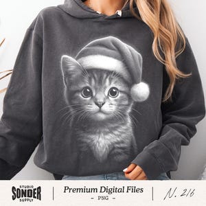 May include: Dark gray hoodie featuring a grayscale image of a kitten wearing a Santa hat. The kitten has large, expressive eyes and a fluffy white pom-pom on the hat. The text "Premium Digital Files" and "Studio Sonder Supply" are also visible.