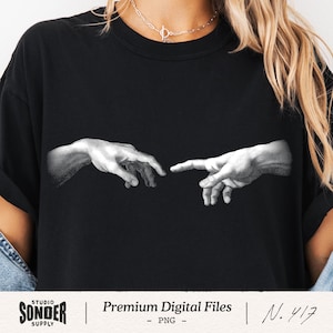 May include: Black t-shirt featuring a grayscale image of two hands nearly touching, a detail from Michelangelo's The Creation of Adam. The shirt includes the text "STUDIO SONDER SUPPLY | Premium Digital Files - PNG - N. 417".