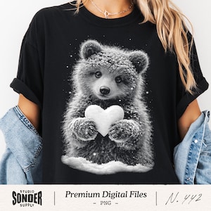 May include: Black t-shirt with a grayscale illustration of a bear cub holding a white heart. The bear is covered in snow, and the design has a winter theme. The shirt has the text "Premium Digital Files" and "N. 442".