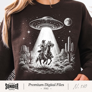 Könnte beinhalten: Braunes Sweatshirt mit einer schwarz-weißen Grafik eines Cowboys auf einem Pferd, der von einem UFO entführt wird. Das Design zeigt eine Wüstenlandschaft mit Kakteen, Bergen, Sternen und einem Mond.