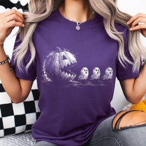 May include: A purple short-sleeved t-shirt featuring a white graphic print of a large, jagged-mouthed jack-o'-lantern chasing three small, wide-eyed ghosts. The design has a distressed, textured appearance, and the shirt has a classic crew neckline.