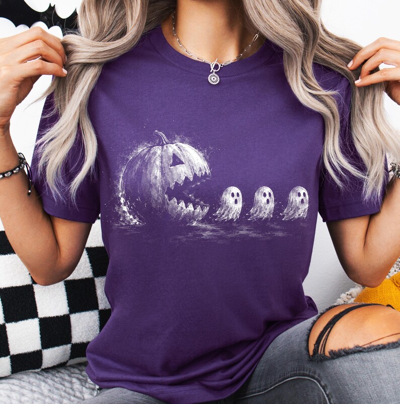 May include: A purple short-sleeved t-shirt featuring a white graphic print of a large, jagged-mouthed jack-o'-lantern chasing three small, wide-eyed ghosts. The design has a distressed, textured appearance, and the shirt has a classic crew neckline.