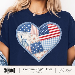Puede incluir: Camiseta azul marino con un diseño de corazón de retazos. El corazón incorpora elementos de la bandera estadounidense, estrellas, rayas y estampados florales. También aparece la Estatua de la Libertad. La camiseta tiene un borde deshilachado.