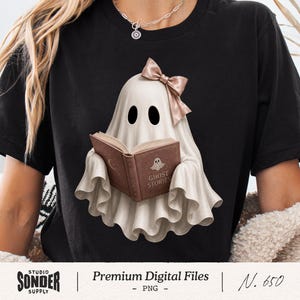 May include: A black t-shirt featuring a graphic of a white ghost wearing a light brown bow and reading a brown book titled Ghost Stories. The ghost has two black oval eyes and is depicted with a draped, sheet-like appearance. The design is centered.