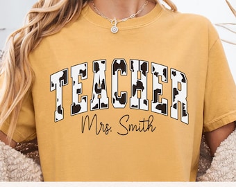 May include: Mustard yellow t-shirt with the word "TEACHER" in a white and black cow print design. Below the word "TEACHER" is the name "Mrs. Smith" in a cursive font. The shirt has a crew neck and short sleeves.