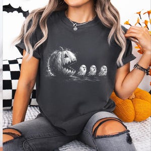 May include: A charcoal grey short-sleeve t-shirt featuring a white graphic print of a large jack-o'-lantern with a wide, jagged mouth chasing three small, white ghosts in a line. The design has a distressed, textured appearance on the fabric.