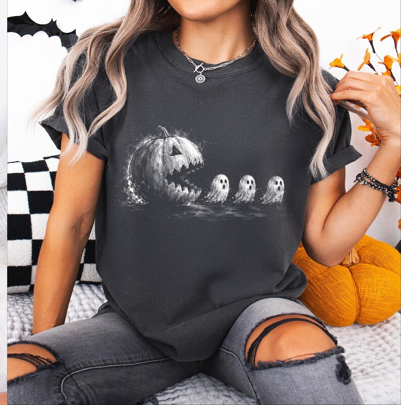 May include: A charcoal grey short-sleeve t-shirt featuring a white graphic print of a large jack-o'-lantern with a wide, jagged mouth chasing three small, white ghosts in a line. The design has a distressed, textured appearance on the fabric.