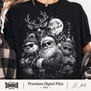 Op de afbeelding: Een zwart t-shirt met een grafische print van de Kerstman, een sneeuwpop, een peperkoekmannetje en een rendier, die allemaal een zonnebril dragen. De scène bevat een volle maan, een silhouet van een arrenslee en een besneeuwd bos.