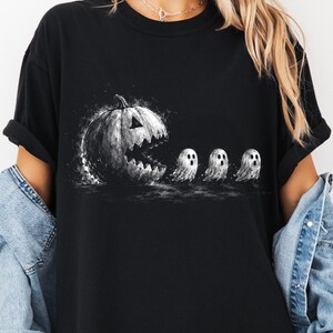 May include: A black short-sleeved t-shirt featuring a graphic print of a large, dark, jagged-mouthed jack-o'-lantern chasing three small, white, wide-eyed ghosts in a stylised, ink-splatter art design across the centre of the chest.