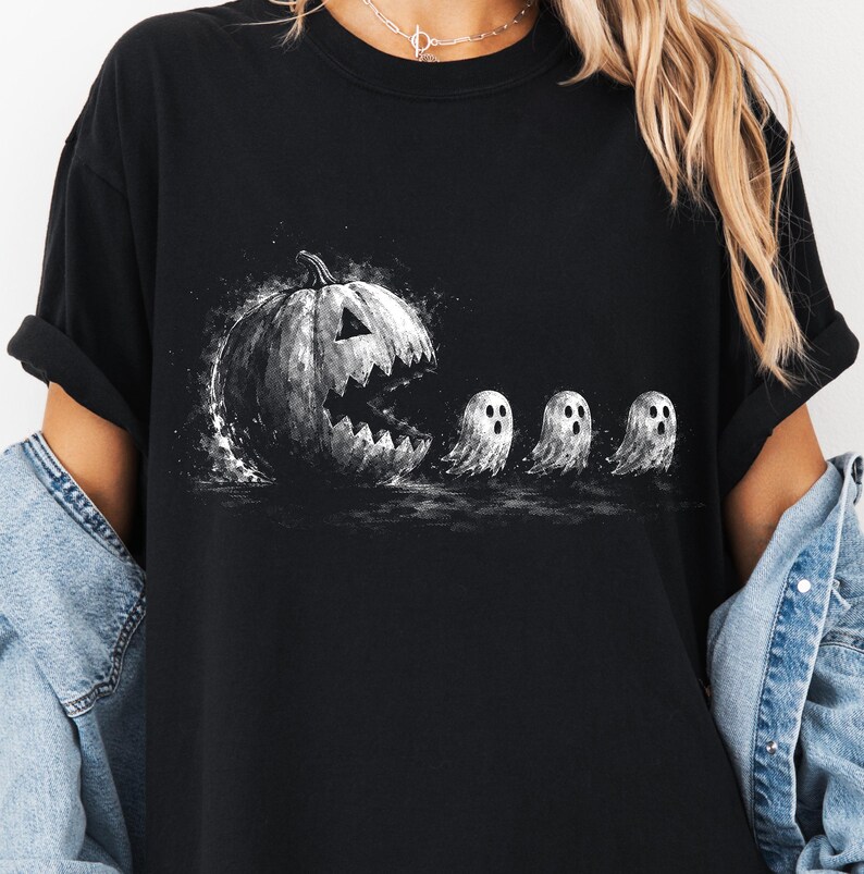 May include: A black short-sleeved t-shirt featuring a graphic print of a large, dark, jagged-mouthed jack-o'-lantern chasing three small, white, wide-eyed ghosts in a stylised, ink-splatter art design across the centre of the chest.