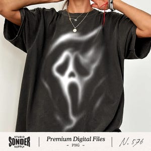 May include: A charcoal black short-sleeve t-shirt featuring a large, blurred, white airbrushed-style graphic of a ghost-like mask with hollow eyes and an elongated mouth centred on the front of the garment, creating a high-contrast visual effect.
