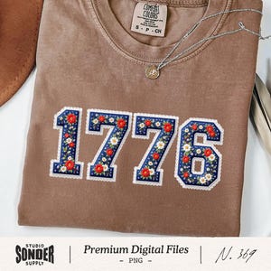 May include: A brown Comfort Colors t-shirt featuring the number "1776" in a floral design. The numbers are blue with red and white flowers and a white lace trim. A silver necklace with a pendant is visible.