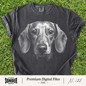 Puede incluir: Una camiseta de manga corta en color gris carbón con un estampado gráfico realista en blanco y negro del rostro de un perro salchicha en el centro. La prenda tiene cuello redondo y un aspecto de tejido lavado vintage.